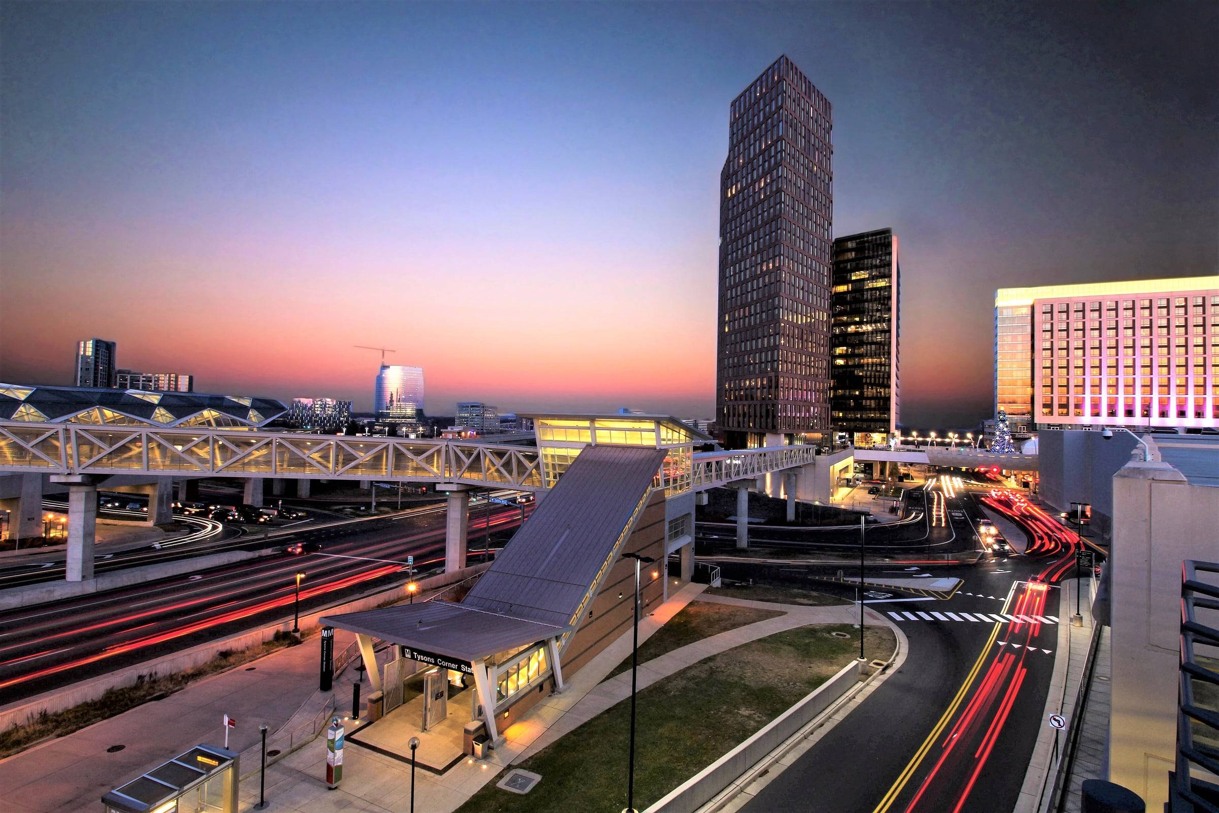 Tysons office towers and the Metro Silver Line station glowing at sunset, with light trails on the road below